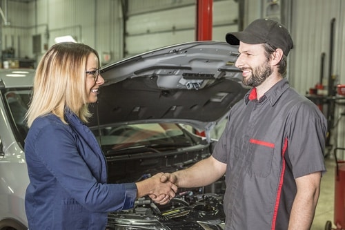 The Importance of Communication from Your Auto Tech with Mast Service Center in Etna Green, IN; image of tech shaking hands with middle aged blonde headed woman in blue blazer in front of her car with hood open in shop bay area