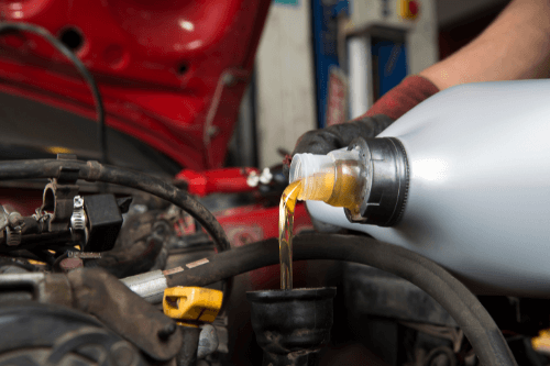 An Inside Look at What Happens to Your Vehicle's Engine Without a Timely Oil Change with Mast Service Center in Etna Green, IN. image of mechanic pouring fresh oil out of large plastic silver oil container on red vehicle in shop