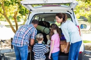 How to Get Your Vehicle Ready for a Road Trip | Mast Service Center in Etna Green, IN. Image of a happy family getting ready for road trip on a sunny day, packing their belongings into their white car.