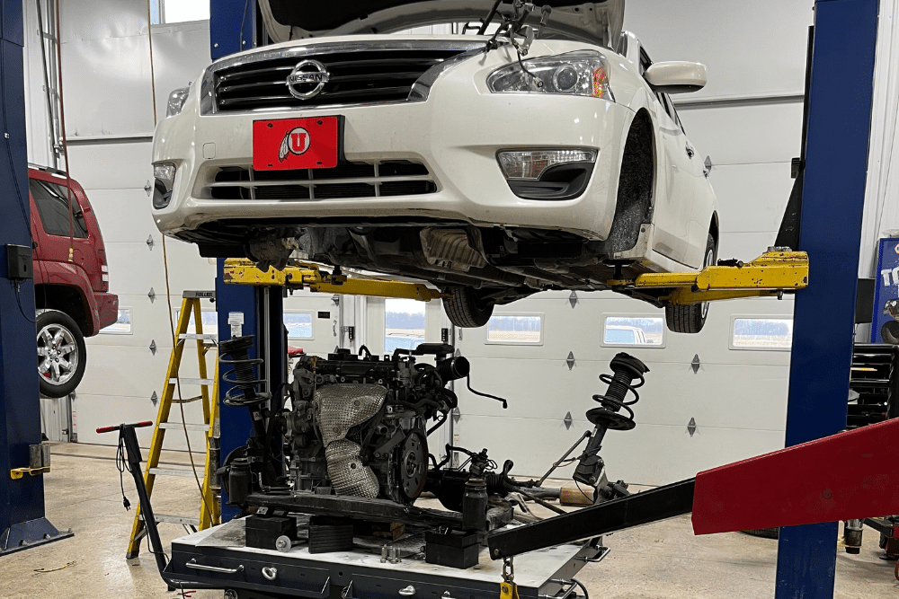 car problems in Napanee, IN at Mast Service Center. Engine and transmission removed from a white Nissan sedan on a lift inside a clean auto repair shop, with the drivetrain components laid out on a cart below.