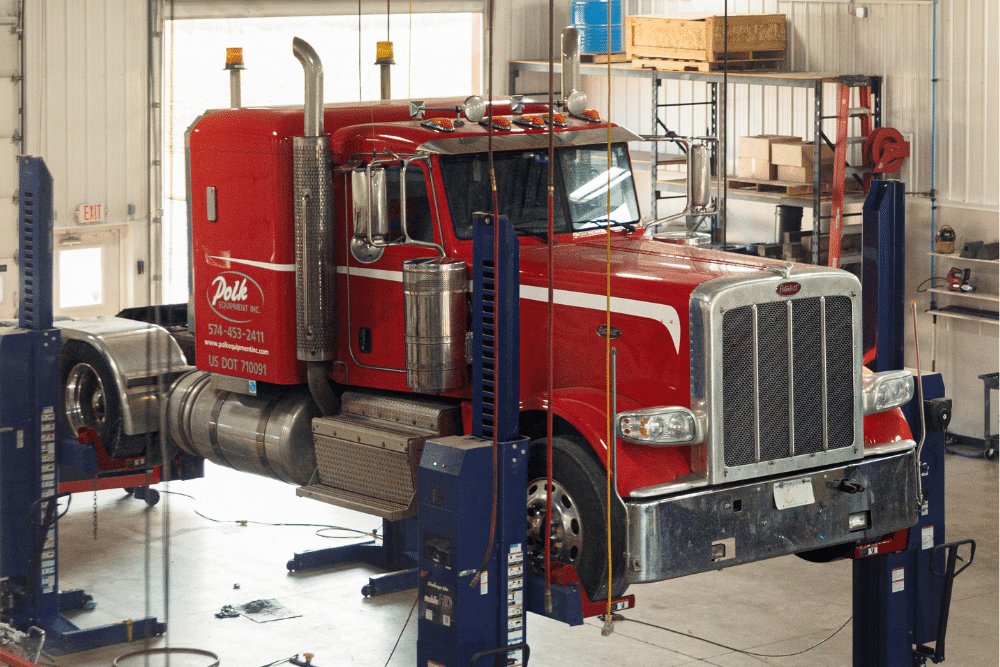 Scheduled Fleet Service in Nappanee, IN at Mast Service Center. Red semi-truck lifted on hydraulic service lifts inside a commercial repair shop for fleet maintenance.